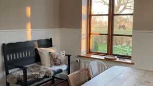 A room with a wooden table and bench, a large window with a view of a field and trees, and a radiator under the window. - Vacation accommodation "Verwalterhaus" with view of the horse pasture -Gut Wittmoldt- (Wittmoldt)