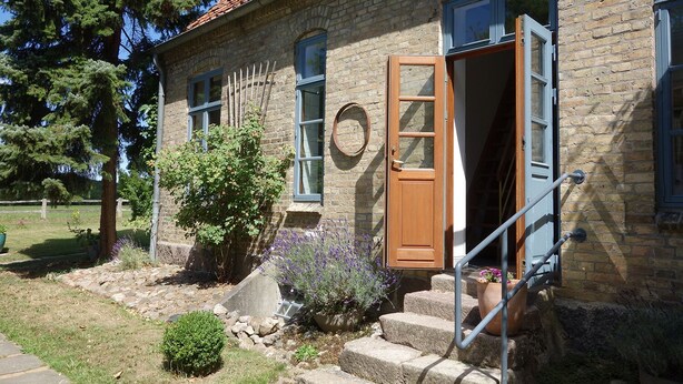 A stone building with a wooden door and windows, a stone staircase, and a garden with plants and a tree. - Vacation accommodation "Verwalterhaus" with view of the horse pasture -Gut Wittmoldt- (Wittmoldt)