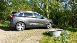 A silver SUV parked on a paved path in a wooded area with a large planter and flowers nearby. - Vacation accommodation "Verwalterhaus" with view of the horse pasture -Gut Wittmoldt- (Wittmoldt)