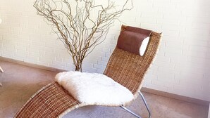 Un fauteuil de relaxation en osier, doté d’un coussin d’assise blanc et moelleux, est placé devant un mur de briques blanches, au-dessus duquel est accrochée une œuvre d’art encadrée.
