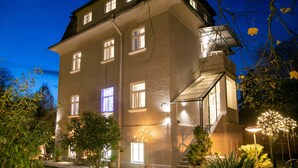 A multi-story building with illuminated windows and outdoor lighting, surrounded by trees and plants.