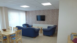 Een kamer met twee blauwe banken, een houten eettafel met stoelen, een aan een bakstenen muur bevestigde televisie en een radiator.