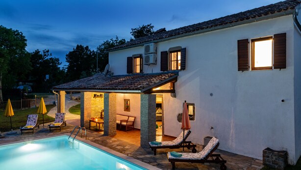 Un área de piscina con sillones de descanso y sombrillas, un patio de piedra y un edificio con tejado de tejas y ventanas.