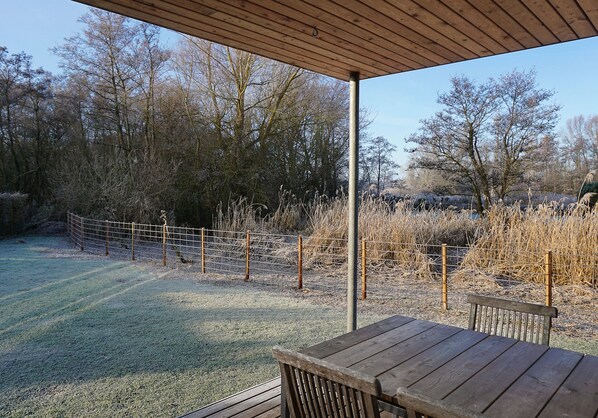 An outdoor wooden deck with a table and bench, surrounded by a fenced area with trees and dry vegetation.