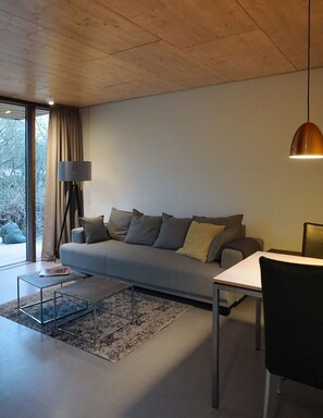 A modern living room with a grey sofa, a small table, a lamp, and a view of trees through a large window. - noise | Design cottage with 2 apartments (Timmendorfer Strand)
