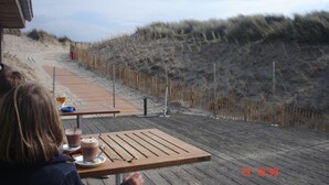 A wooden table with a drink and a plate, overlooking a sandy beach with a fence. - Bright vacation home for up to 6 people with fireplace, garden, parking and WiFi in De Haan (De Haan)