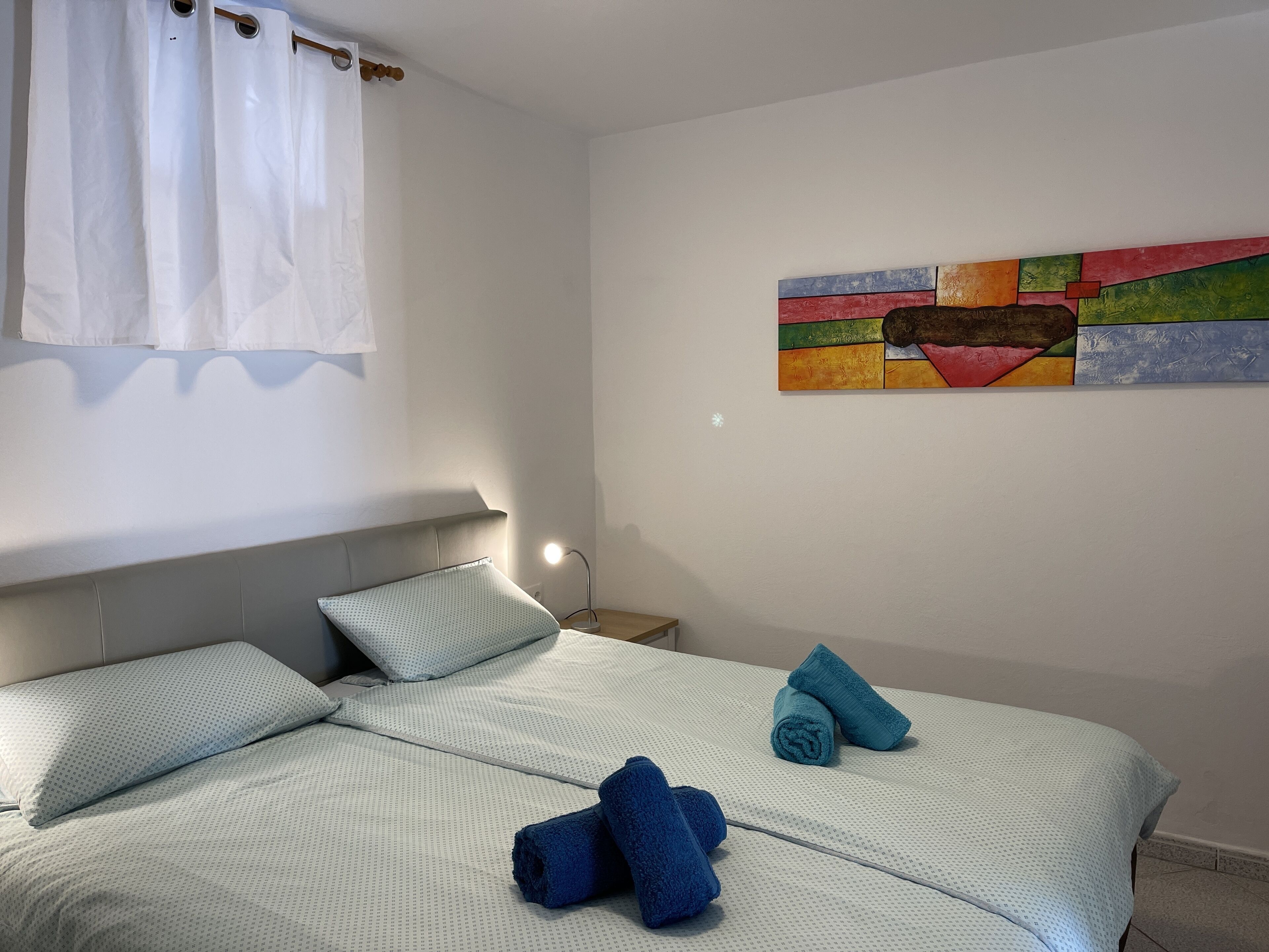 A hotel room with a bed, pillows, a nightstand, a lamp, and a colorful abstract painting on the wall.