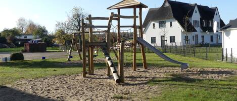 Un parque infantil con tobogán y estructuras para escalar, rodeado de arena y césped, con una casa al fondo.