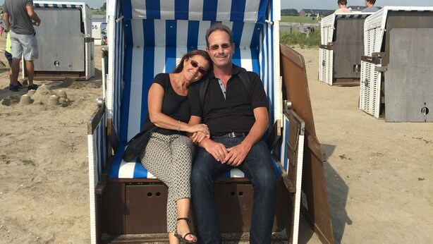 Una pareja sentada en una silla de playa a rayas sobre arena, con baños portátiles al fondo.