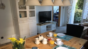 Una mesa de comedor dispuesta con platos, tazas y una jarra, con un televisor al fondo y vista a un jardín por una ventana.