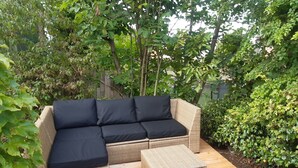 A wicker sofa with black cushions and a matching ottoman on a wooden deck surrounded by lush greenery.
