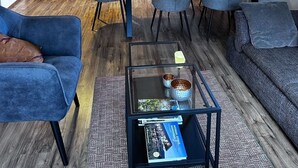 A modern kitchen with wooden beams, a dining area with blue chairs, and a living room with a sofa and a coffee table.