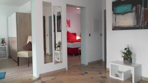A modern living room with a sofa, a coffee table, and a mirror. There is a hallway leading to a bedroom with a red bed and a painting on the wall. - Welcome Holiday home Schmitz No. 2, for 2 to 4 people.  with Wi-Fi (Pünderich/Mosel)