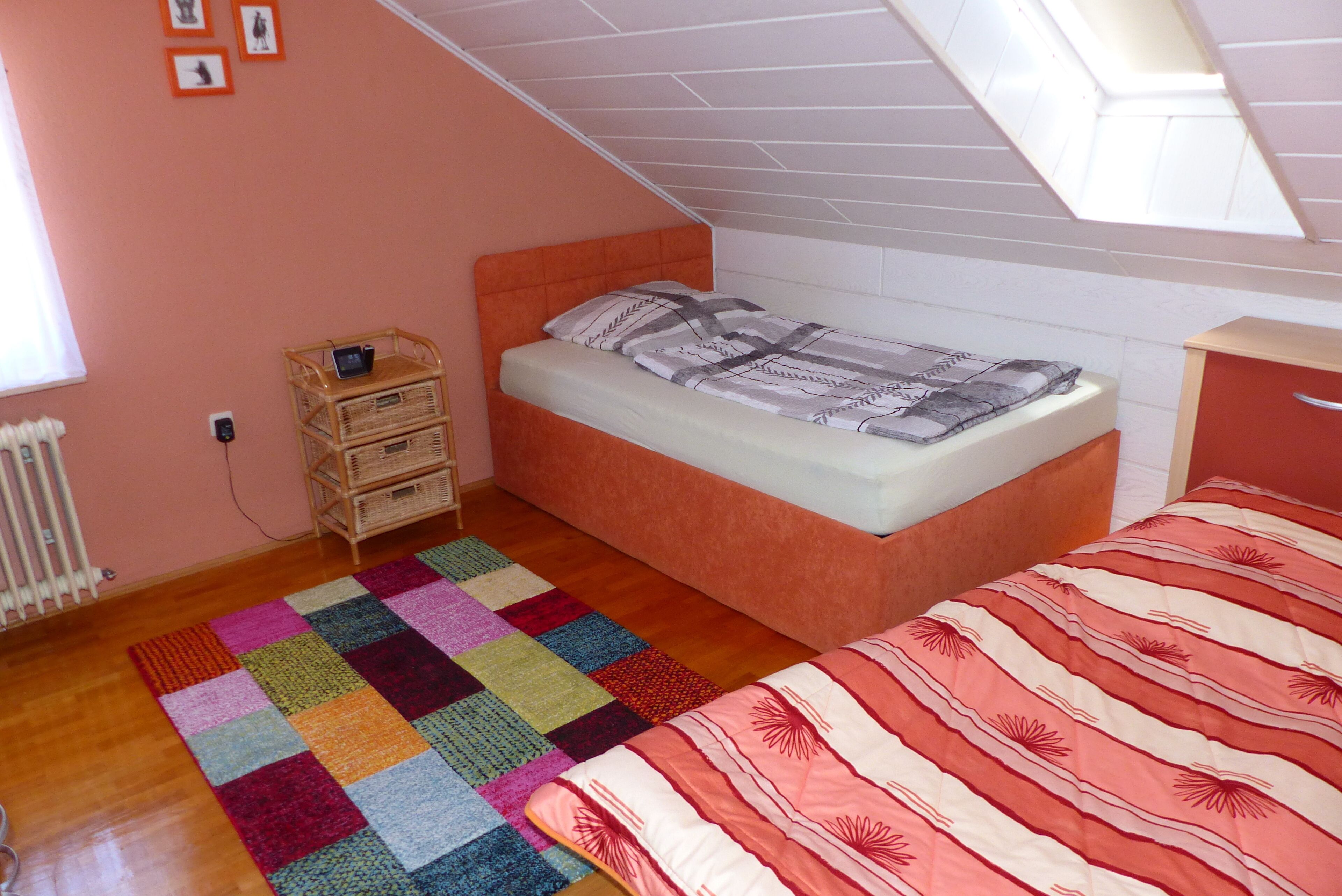 A bedroom with a single bed, a colorful rug, a small wooden side table, and a window with blinds.