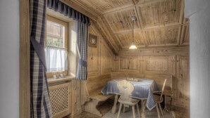 Une pièce chaleureuse aux murs et au plafond en bois, une fenêtre avec des rideaux, une table recouverte d’une nappe à carreaux et une seule chaise.