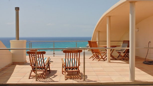 Una terraza con sillas y una mesa de madera, con vista al mar azul y despejado.