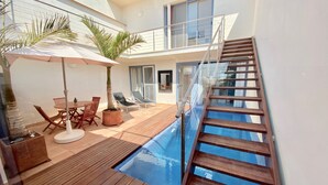 Un área de piscina moderna al aire libre con cubierta de madera, sillones de descanso y una escalera que lleva a un nivel superior.