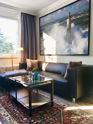 A modern living room with a black sofa, a glass coffee table, and a large painting of a lighthouse on the wall.