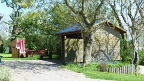 A rustic wooden cabin with a covered porch, a swing set, and a picnic table in a lush green setting.