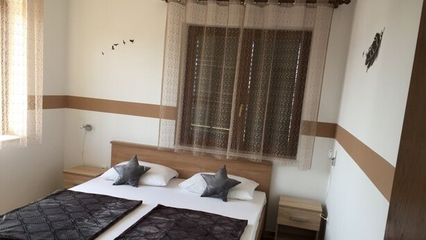 Un dormitorio con cama, dos almohadas, una mesita de noche, una ventana con cortina y una puerta.