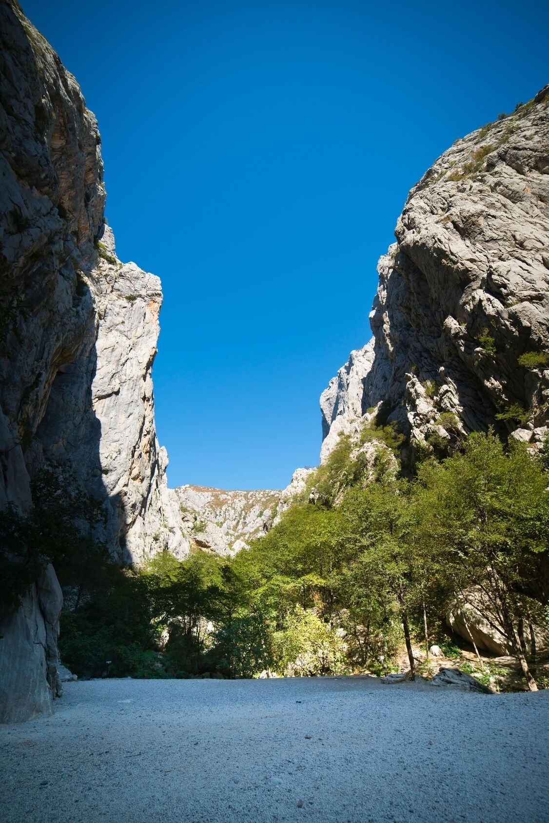 A rocky landscape with a clear blue sky, featuring a gravel path leading through a narrow canyon flanked by steep rock walls and green vegetation.