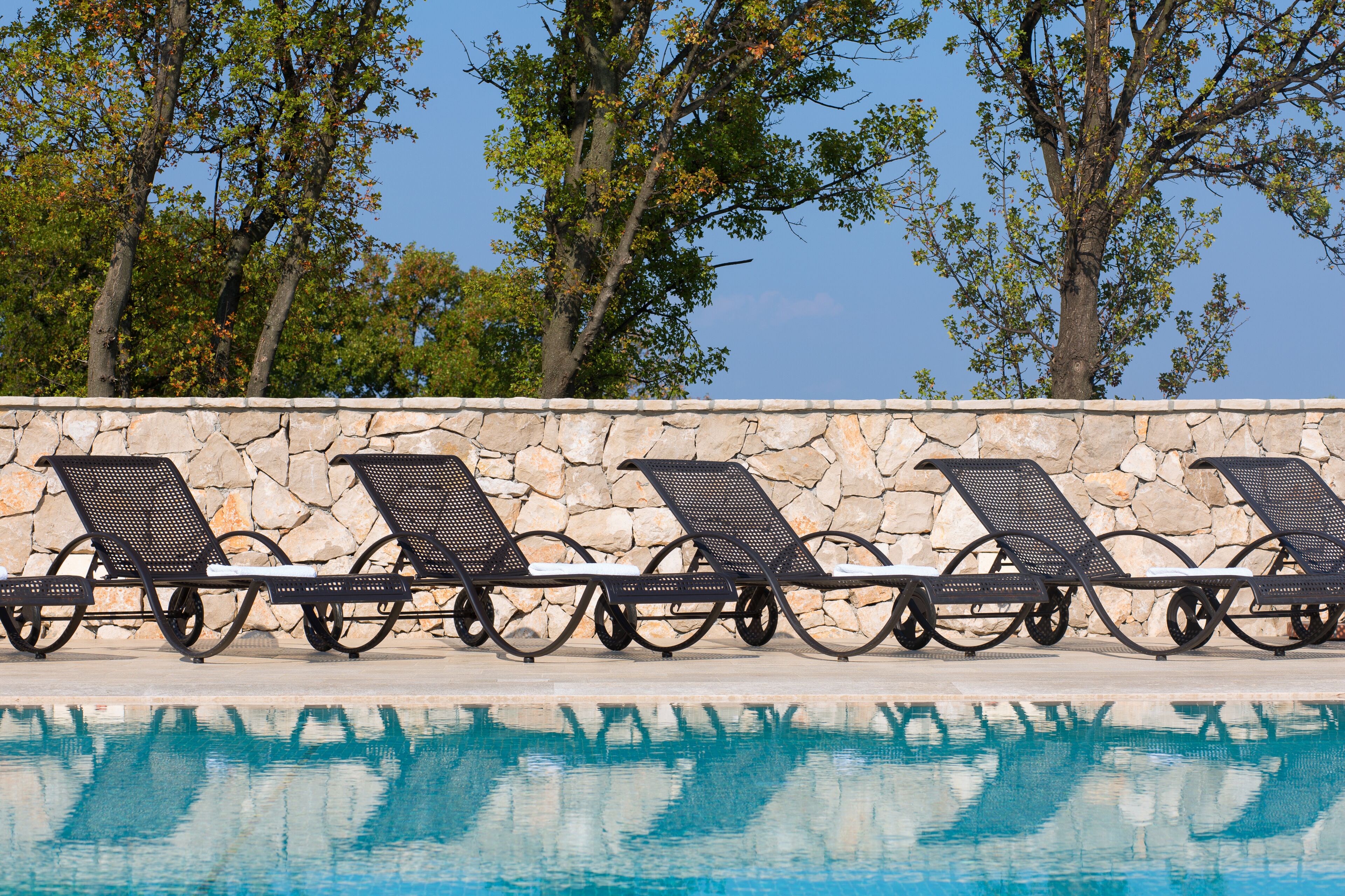 A poolside area with lounge chairs aligned against a stone wall, overlooking a clear blue pool.