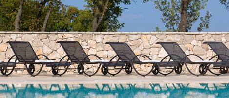 A poolside area with lounge chairs aligned against a stone wall, overlooking a clear blue pool.