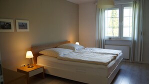 Uma cama bem arrumada com cabeceira de madeira, uma mesa de cabeceira com abajur e uma janela com cortinas.