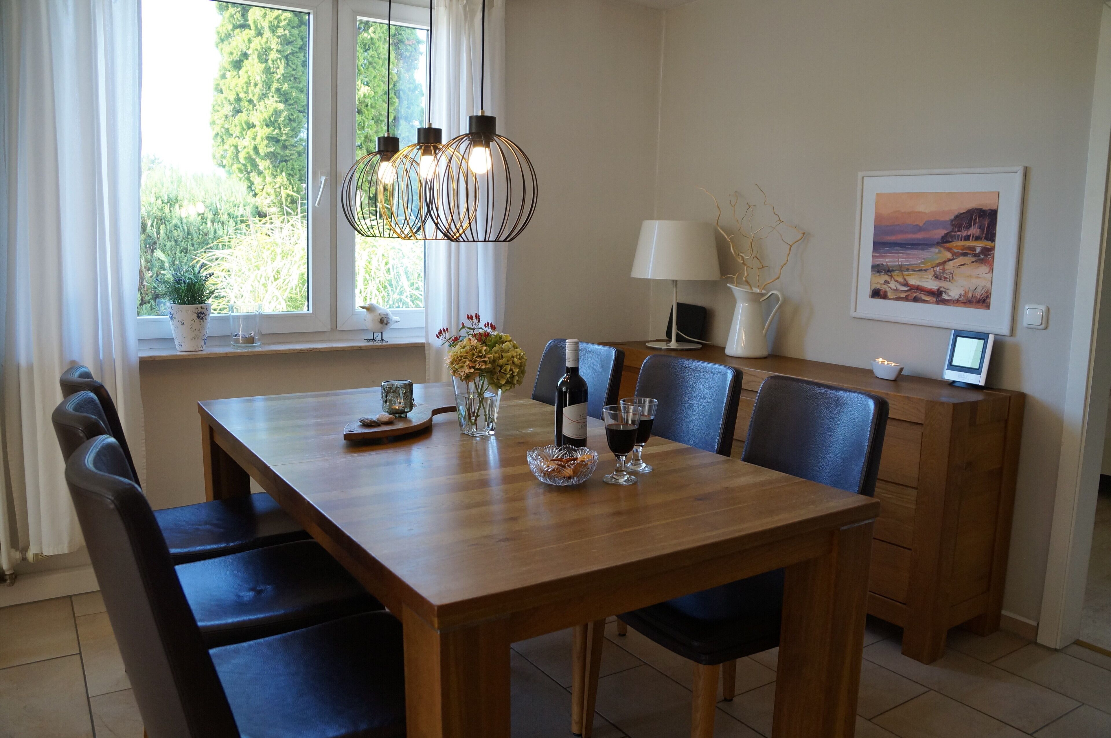 Une salle à manger avec une table en bois, des chaises, un lustre, un vase de fleurs, une bouteille de vin et un tableau encadré accroché au mur.