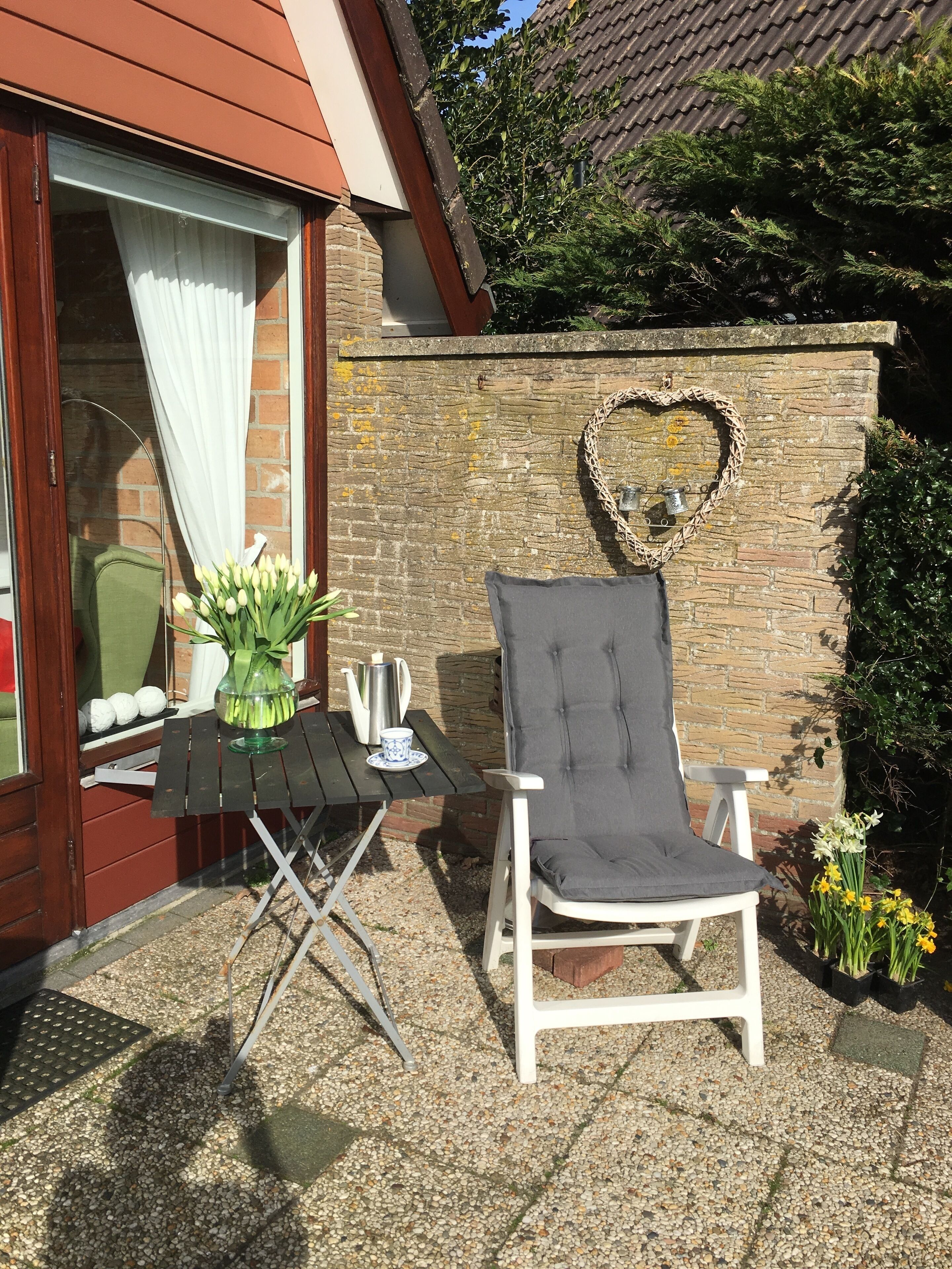 Een terras met een tafel gedekt voor twee personen, een witte stoel en een hartvormige decoratie aan een stenen muur.