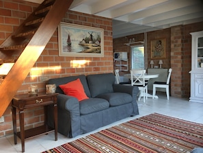 A cozy living room with a brick wall, a wooden staircase, a gray sofa with a red pillow, a white table, and a patterned rug.