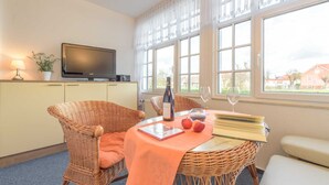 Un comedor con una mesa de mimbre, un televisor sobre un mueble, una botella de vino y vistas al exterior a través de amplias ventanas.
