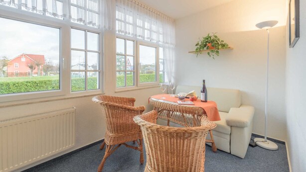 Una habitación con una mesa preparada para dos personas, sillas de mimbre, un sofá blanco y vistas a una casa roja a través de amplios ventanales.