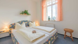 Cama doble con sábanas blancas, mesita de noche con lámpara, ventana con cortinas translúcidas y un estante con una planta.