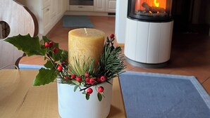 Une pièce chaleureuse avec un décor intérieur en bois, une cheminée centrale, une table à manger agrémentée d’une bougie et de quelques plantes, et une cuisine en arrière-plan.