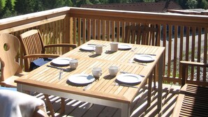 Une terrasse en bois avec une table dressée pour le repas, offrant une vue sur un paysage montagneux avec des champs verdoyants et des bâtiments dispersés.