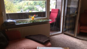 A cozy room with a sofa, a table with a plate, a red chair, and a view of a village through a window.