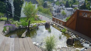 A wooden deck with a pond, surrounded by rocks and plants, overlooking a hillside of houses.