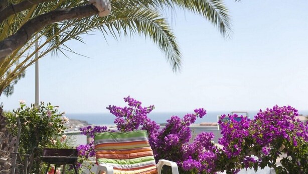 Ein Liegestuhl am Strand mit gestreifter Auflage, umgeben von violetten Bougainvillea-Blüten, mit Blick auf die Küste.