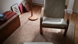 Ein gemütliches Zimmer mit einem Holzstuhl und Hocker, einem Heizkörper, einer Stehlampe und Blick ins Freie durch große Fenster.