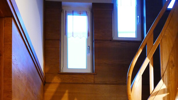 Eine Holztreppe mit Handlauf, die zu einer Zwischenebene mit einem kleinen Fenster führt.