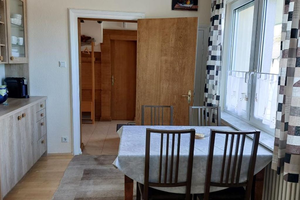Un coin repas avec une table et des chaises, une cuisine équipée d’armoires et d’une machine à café, et une fenêtre avec des rideaux.