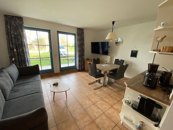 Una sala de estar con sofá, mesa de centro, mesa de comedor con sillas, televisor y área de cocina con cafetera.