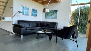 Ein modernes Wohnzimmer mit einer schwarzen Ledersofa, einem Glastisch und einem schwarzen Sessel. An der Wand hängen zwei gerahmte Bilder, eine Stehlampe und ein großes Fenster mit Blick auf Grünflächen.