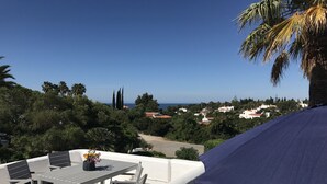 A terrace with a table and chairs, overlooking a coastal town with houses and trees.