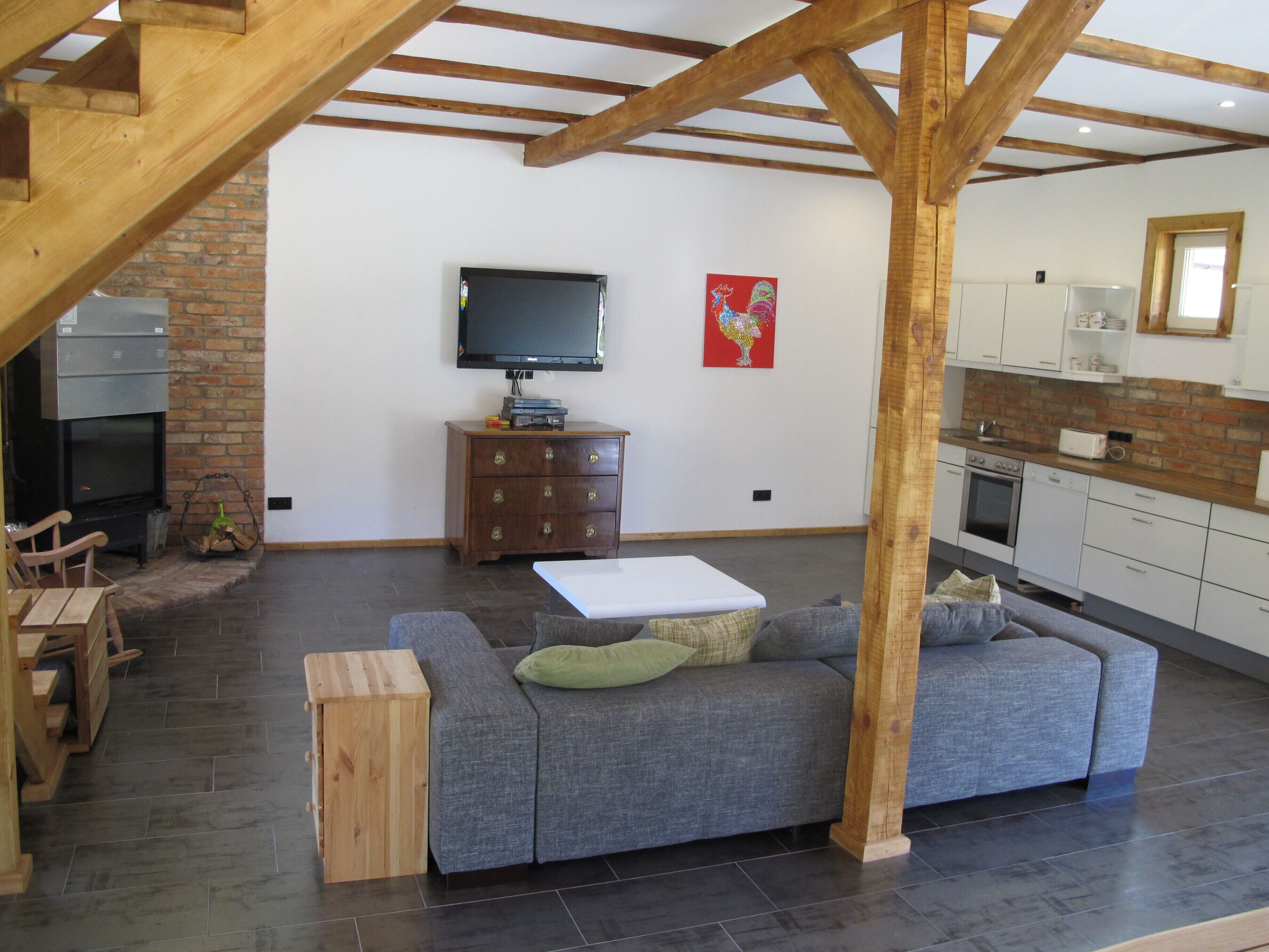 Un vaste espace de vie comprenant un canapé gris, une table basse en bois et un téléviseur fixé au mur. On aperçoit également une cheminée en briques et une cuisine avec des meubles blancs à l’arrière-plan.