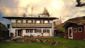 A two-story chalet with a wooden balcony, a stone wall in front, and a small red building to the right. - 80 sqm apartment, 2 bedrooms to 5 pers., 2 balconies, sunny & quiet (Berchtesgaden)