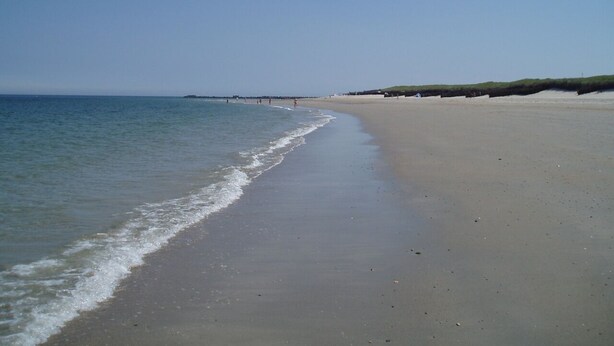 Ein weiter Sandstrand mit sanften Wellen, wolkenlosem Himmel und weiter Bebauung im Hintergrund.