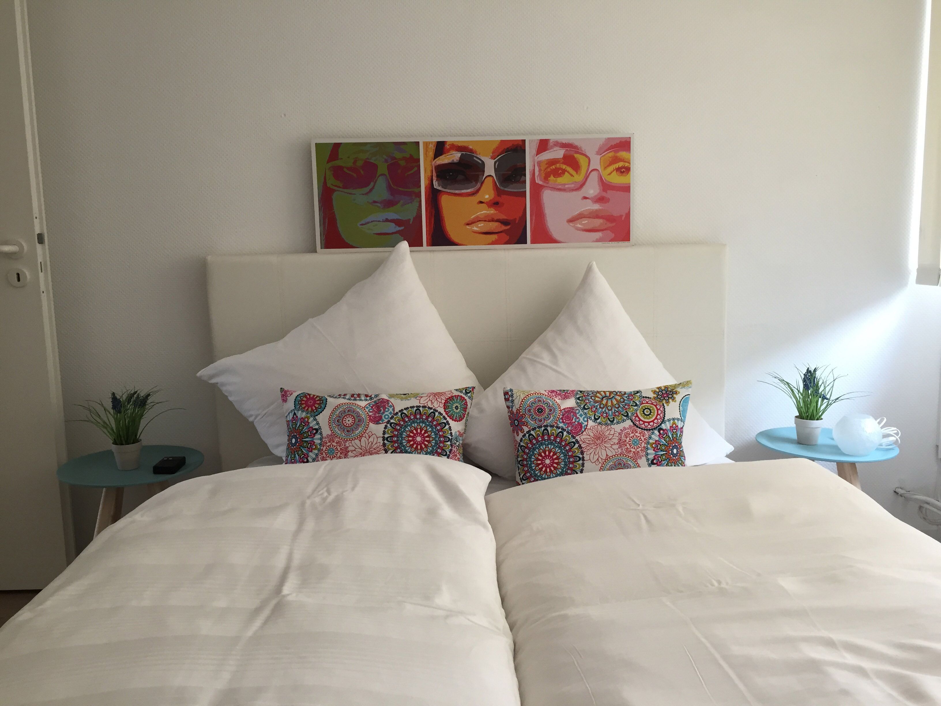 Uma cama com roupa de cama branca, dois travesseiros decorativos com estampas coloridas, e uma parede com três obras de arte emolduradas acima da cama.