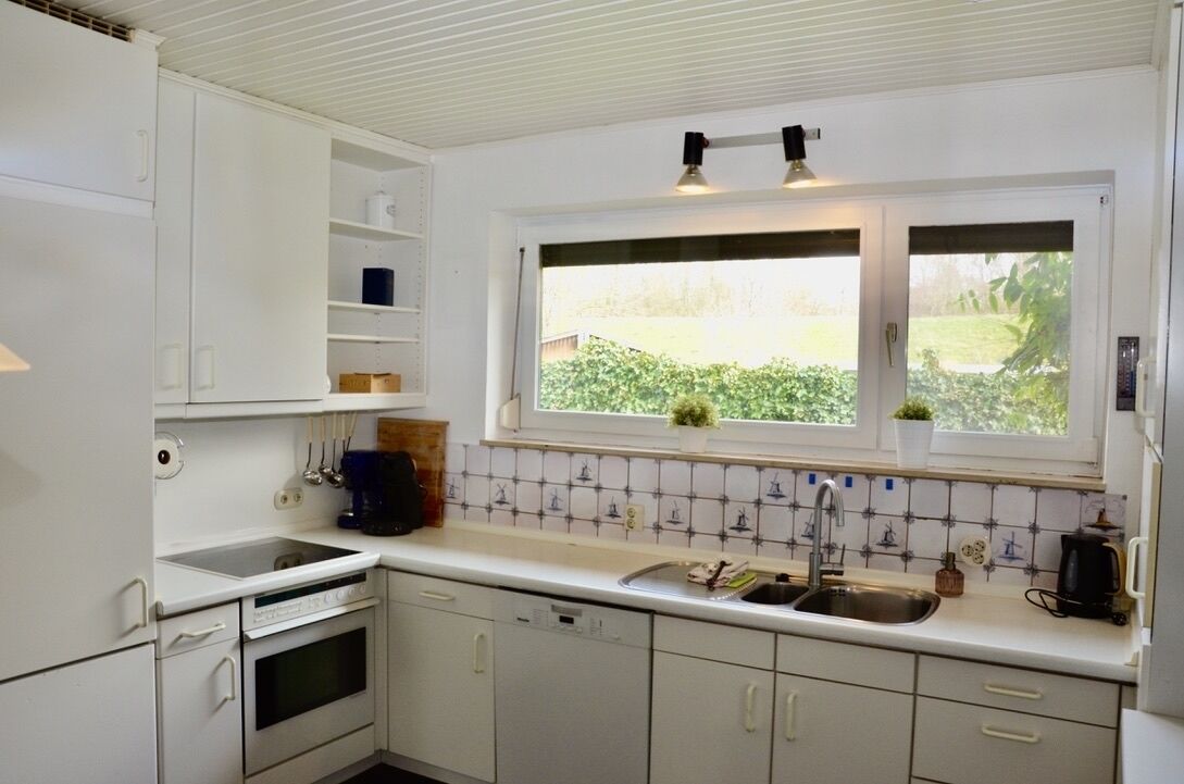 Una cocina con armarios blancos, un lavamanos doble, una ventana con vistas a zonas verdes y un salpicadero de azulejos.